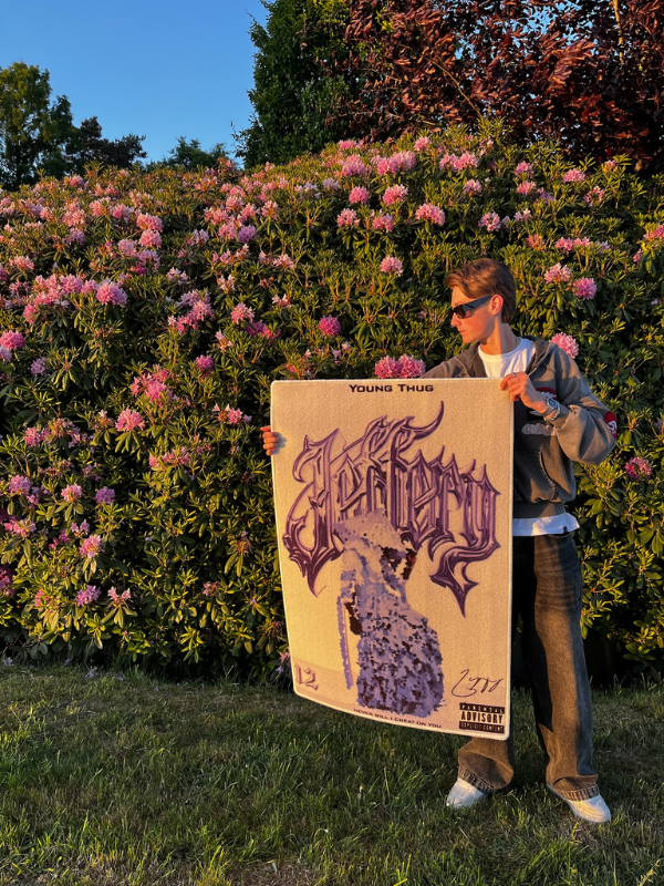 JEFFERY rug – handheld by hedge JEFFERY rug held beside a hedge with purple flowers; white/purple palette complements the scene