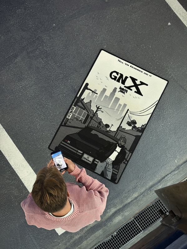 GNX rug – overhead with phone Top-down shot of the GNX rug as a person in a pink sweater photographs it; phone screen visible